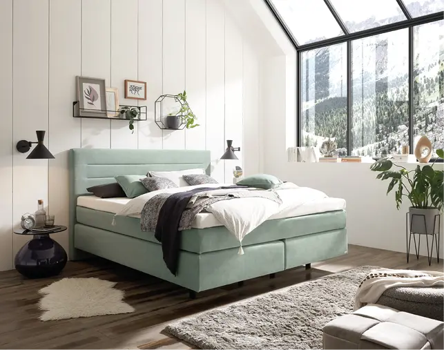 Grünes Polsterbett mit Kissen und Decke vor großer Fensterfront, Teppich und Pflanzen im Schlafzimmer